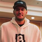 A man standing against a wood paneled wall in a white hooded sweatshirt featuring the Bandits logo and a black Bandits Gaming snapback cap.